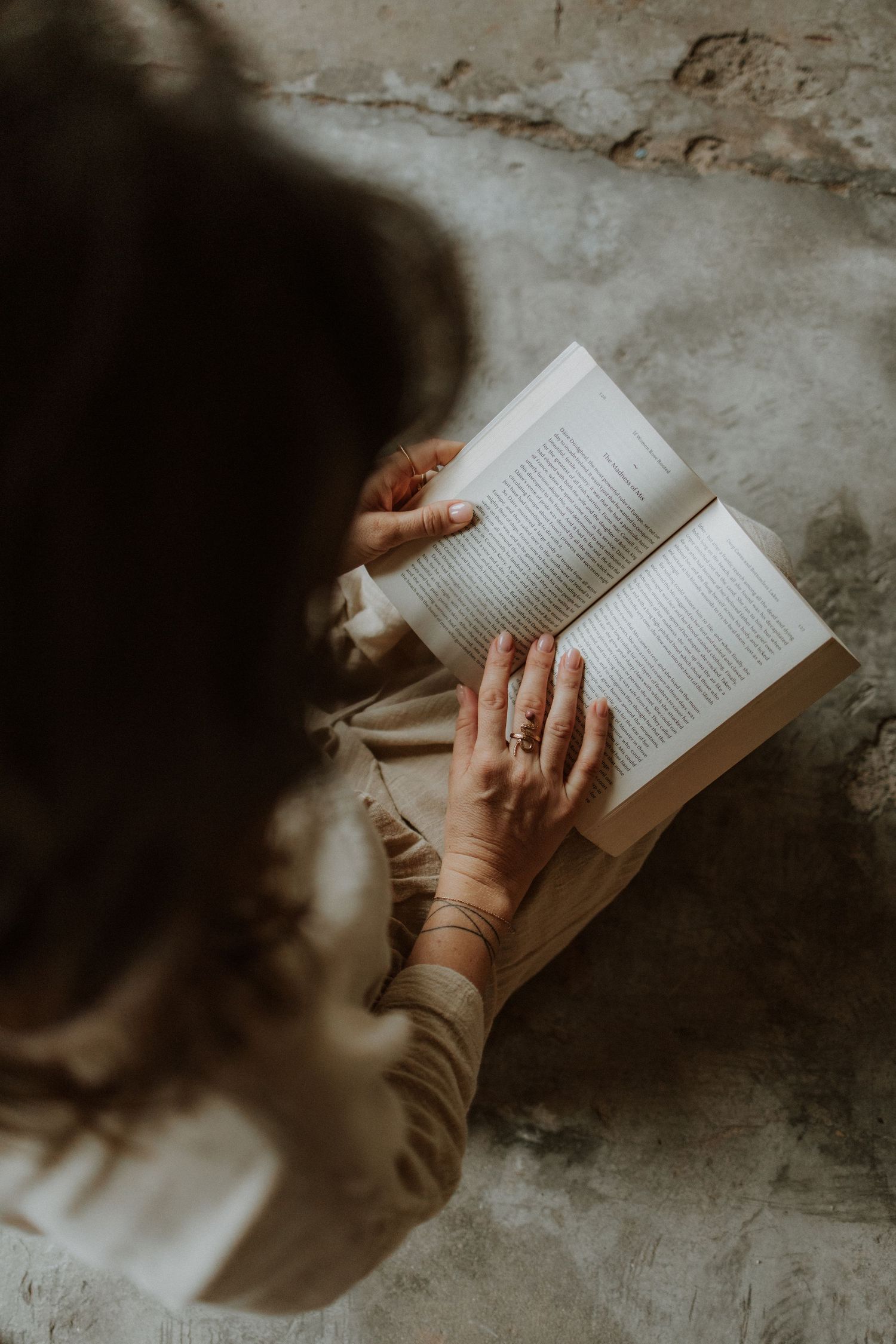A woman with dark hair reads an open book, her ringed hands resting on the pages in a moody, rustic setting.