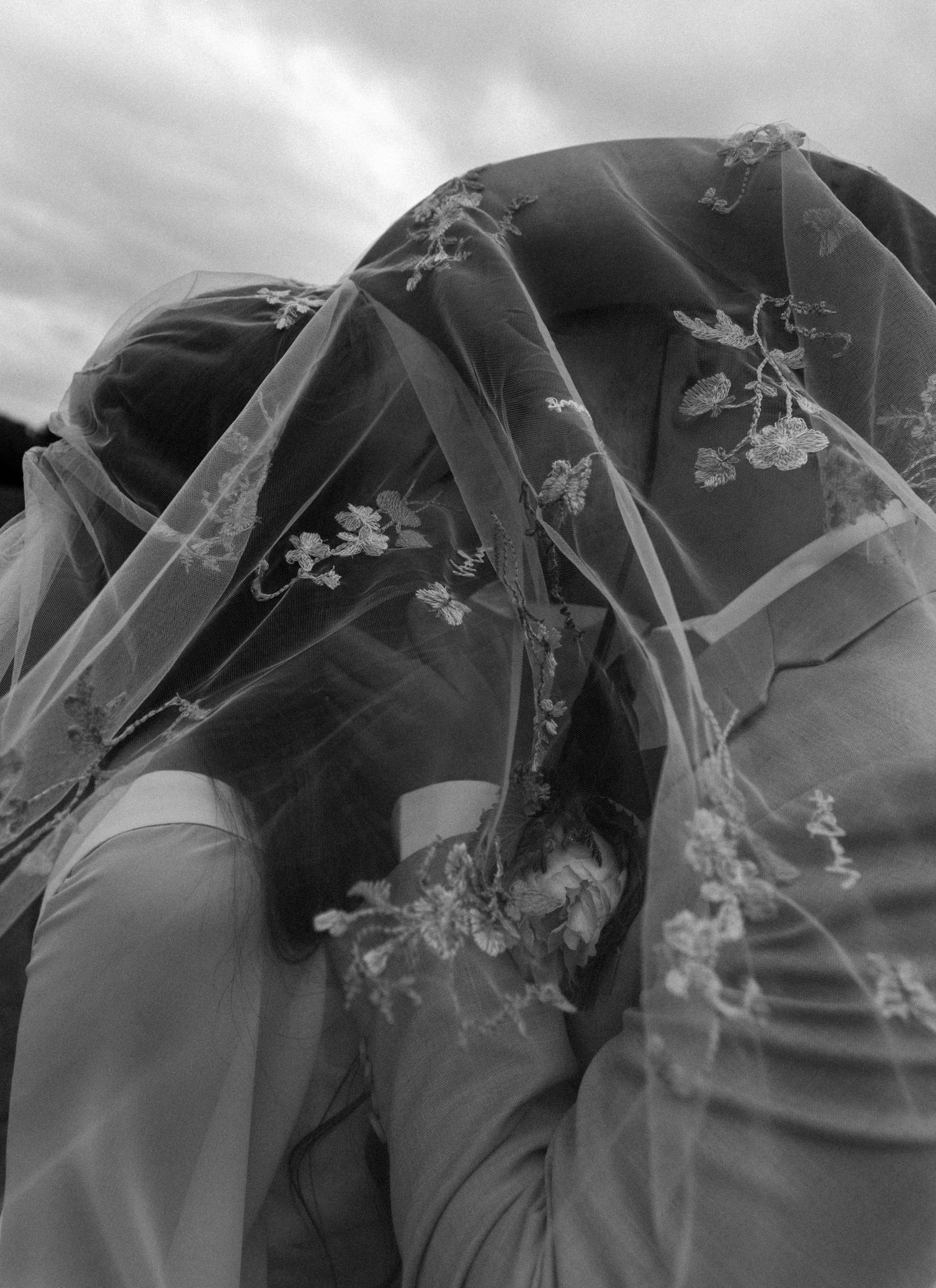 Black and white photo of a bride and groom kissing under a floral embroidered wedding veil outdoors.