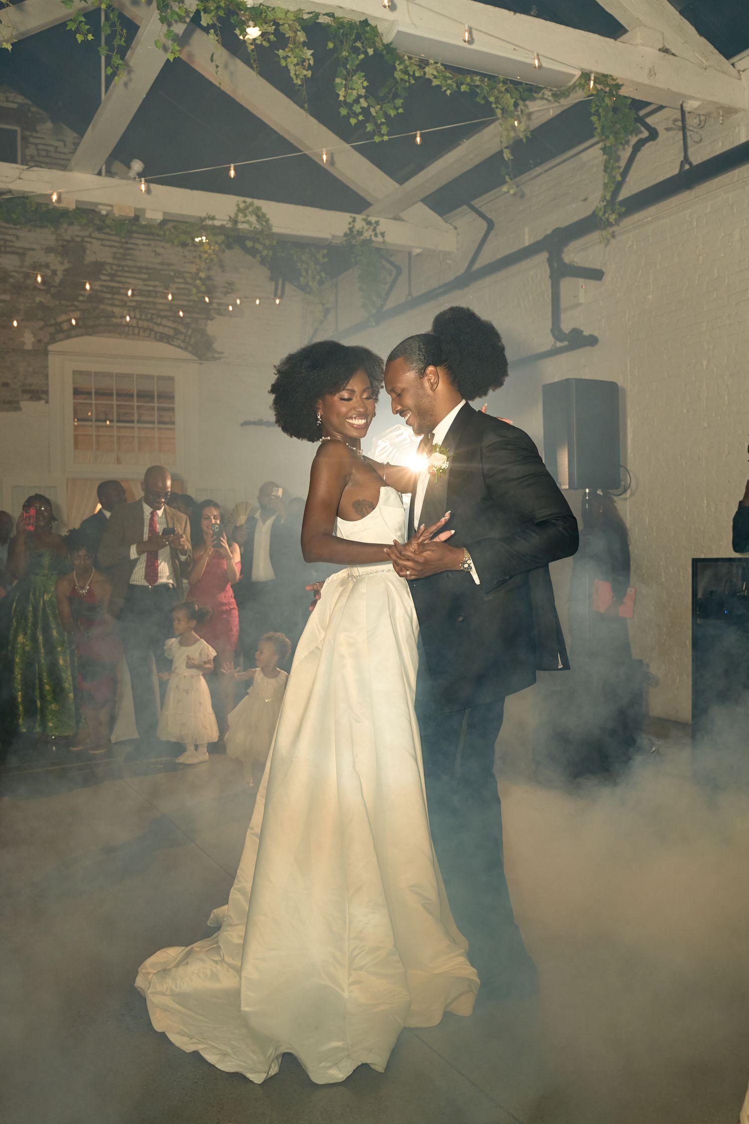 Couple shares first dance surrounded by fog machine smoke at elegant indoor wedding reception with string lights.
