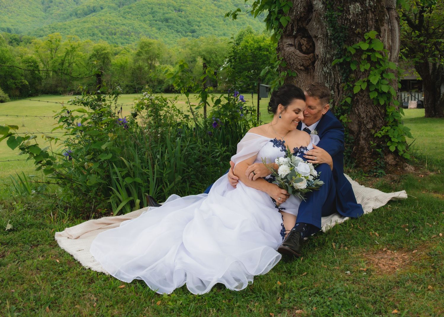 Bride and groom sit together under a tree in a lush green outdoor wedding setting, sharing a romantic moment.