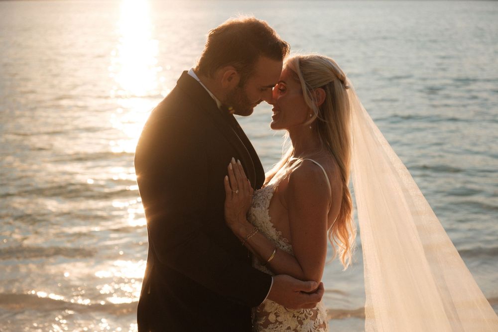 Bride and groom share romantic sunset moment by ocean, her veil flowing as they lean in for a kiss.