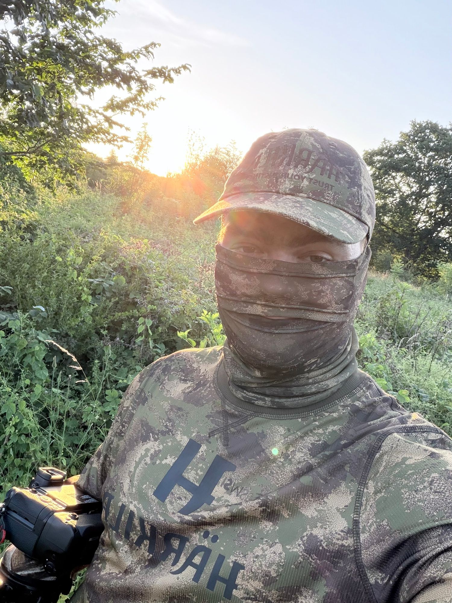 Hunter wearing camouflage clothing and face covering photographed at golden hour outdoors.