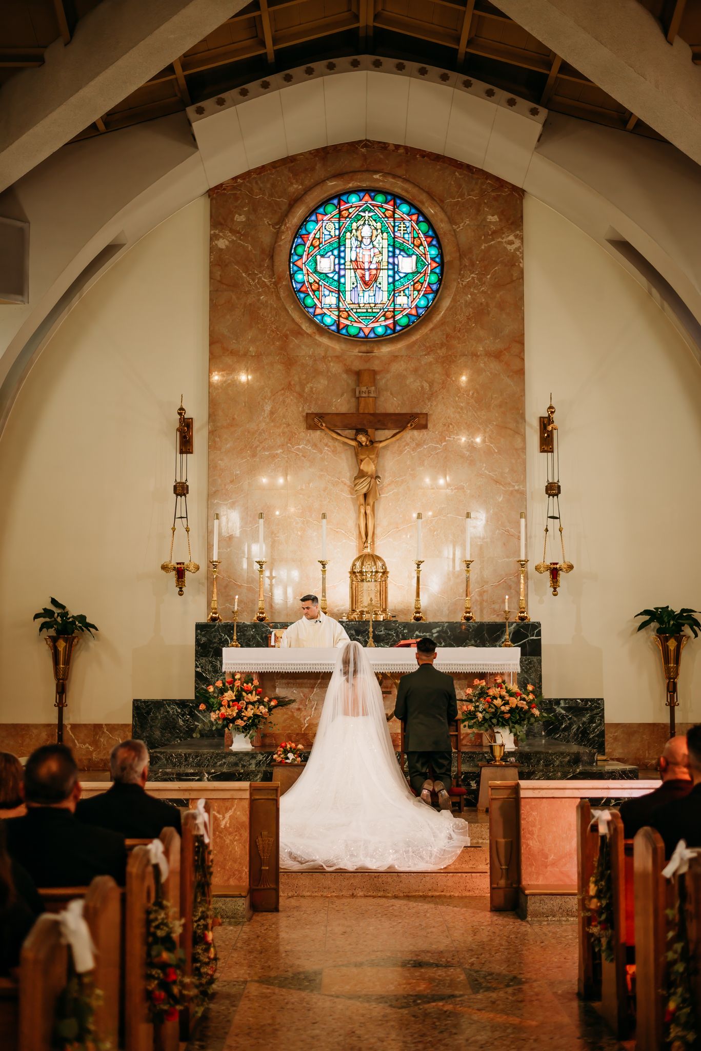 Bride and groom embracing in front of a white gazebo on lush green grass, bride wearing a lace mermaid gown with veil.