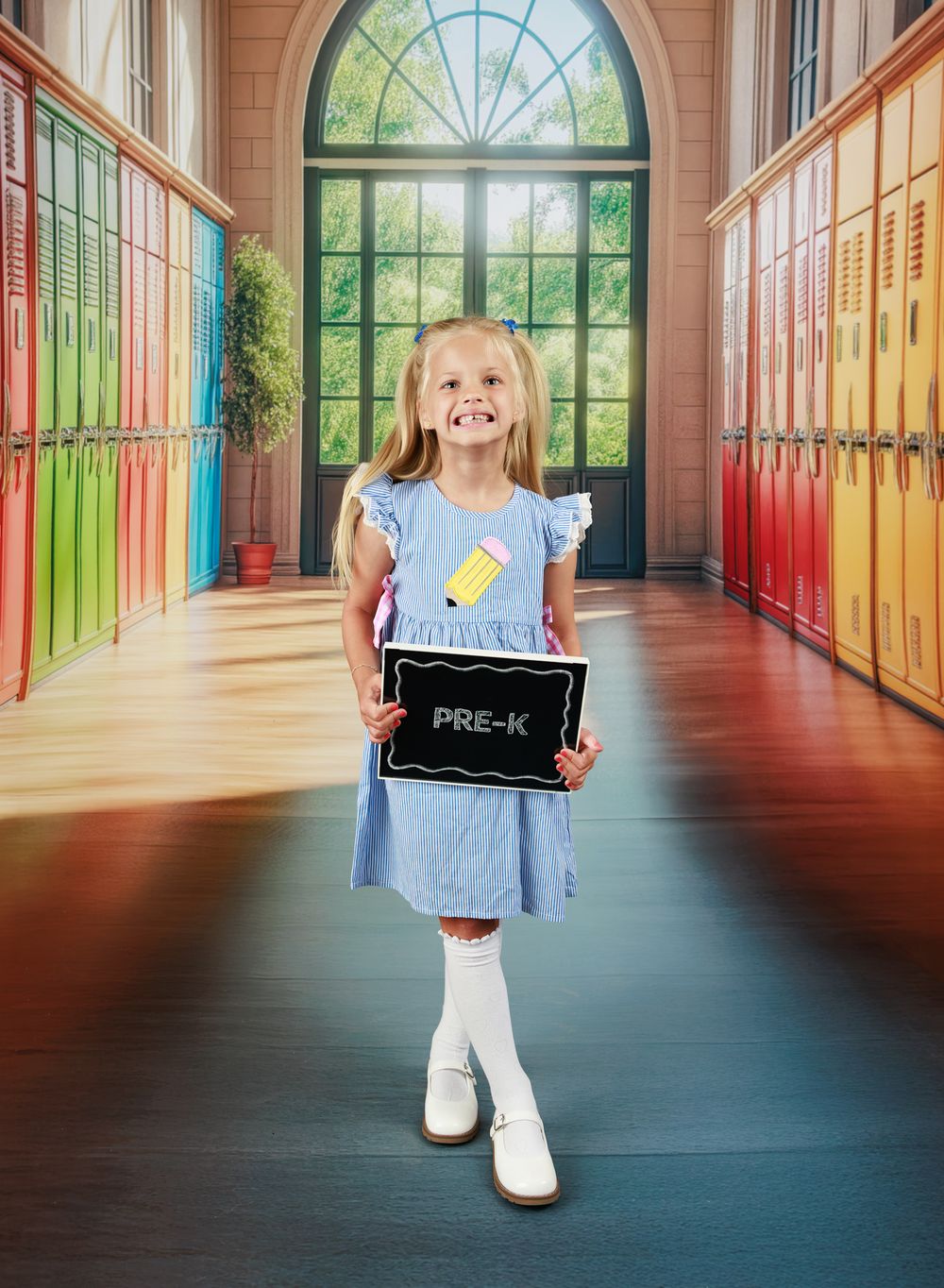 A smiling girl in a blue dress holds a Pre-K chalkboard sign in a colorful school hallway with lockers.