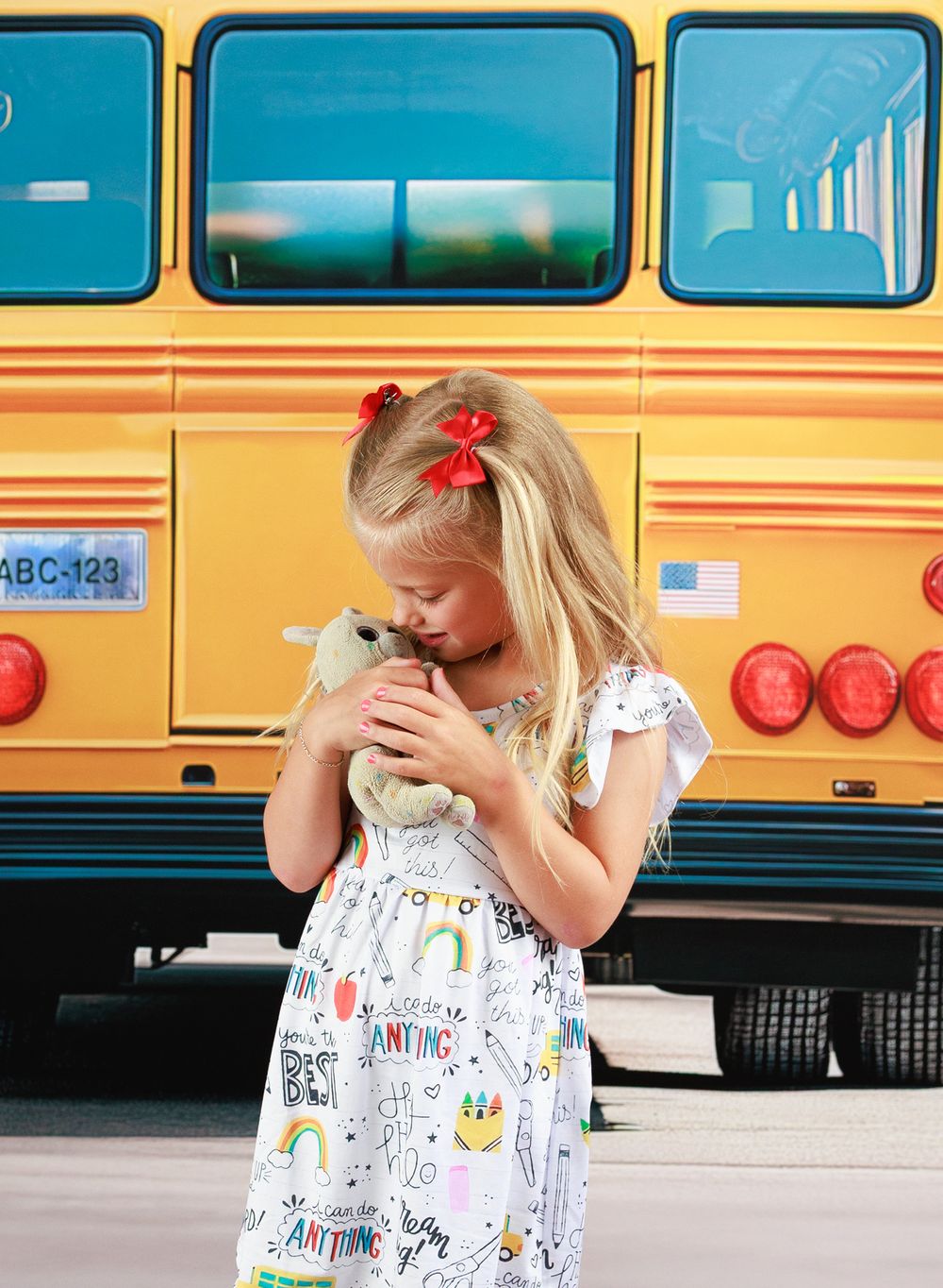 Young blonde girl in a colorful dress hugging a stuffed animal in front of a yellow school bus.