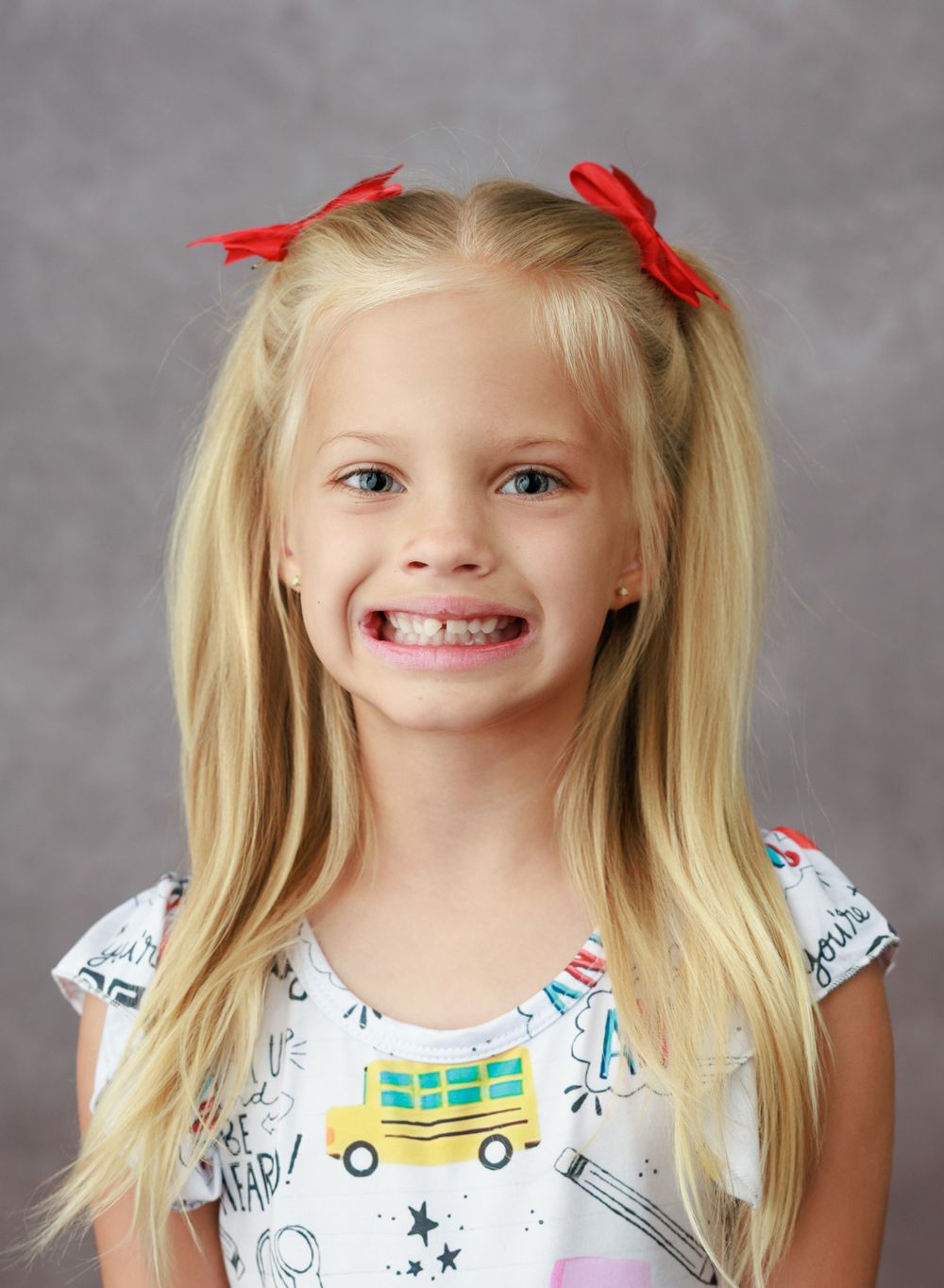 Smiling blonde girl with red hair bows wearing a school-themed shirt, perfect for back-to-school portrait photography.