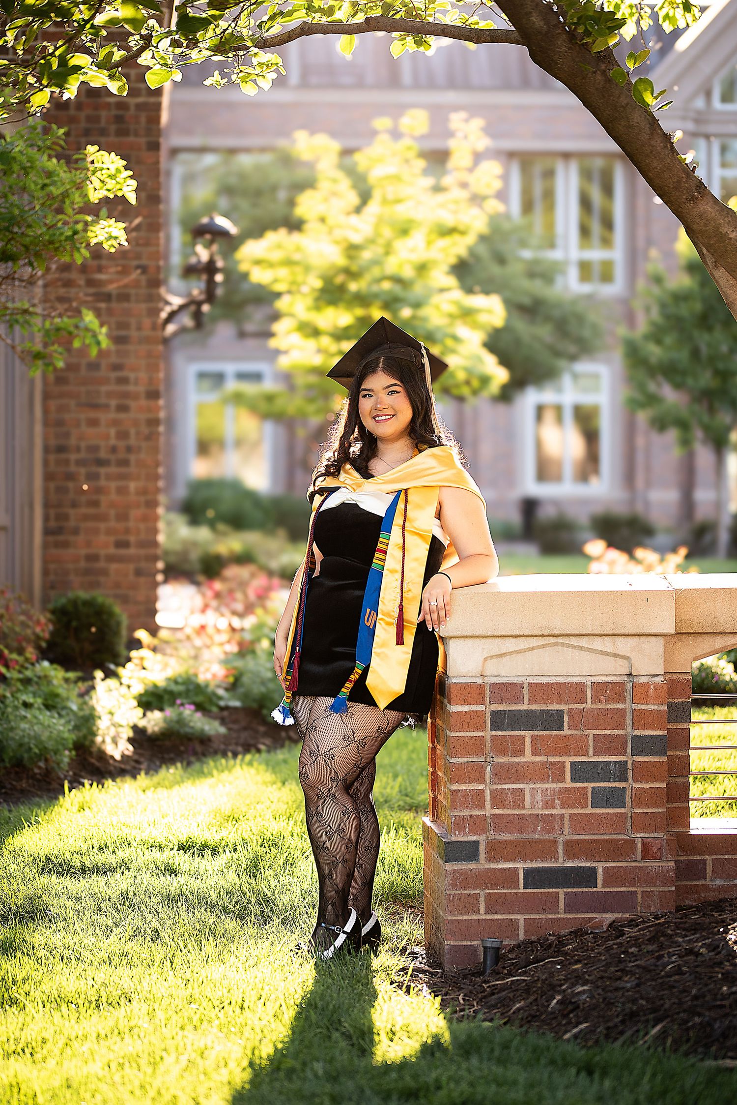 Graduate in black gown with gold stole leaning on brick pillar on sunny campus.