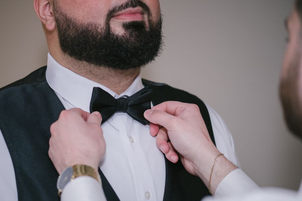 A bearded groom in a black vest and white shirt has his black bow tie adjusted by another person.