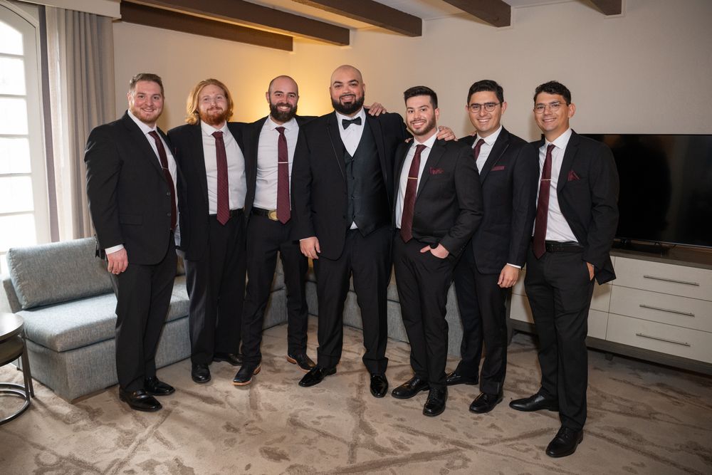 Seven men in black suits with burgundy ties and pocket squares posing together in an elegant room.