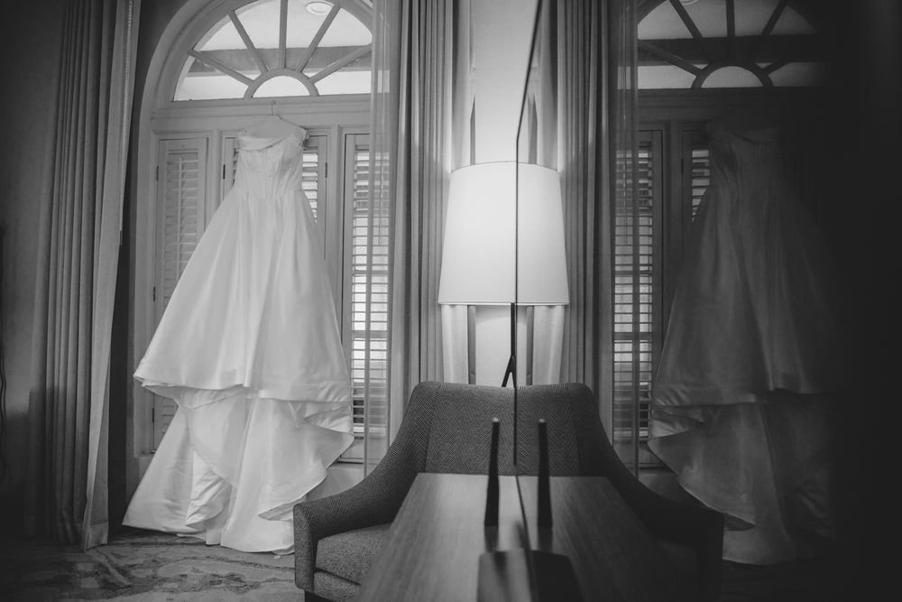 Black and white photo of an elegant wedding dress hanging near a window with shutters and a floor lamp.