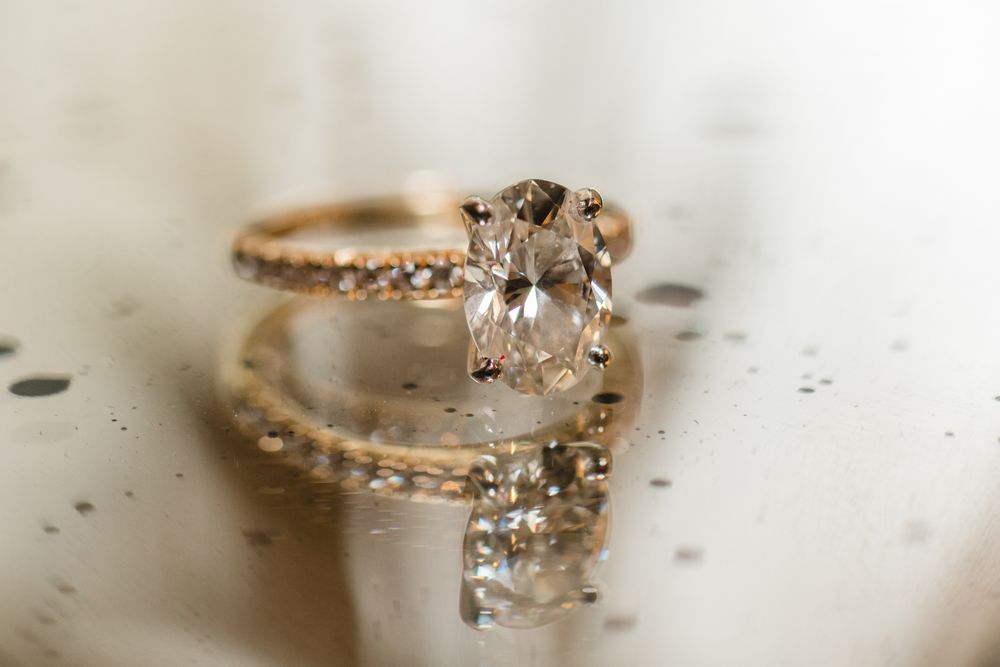 Gold engagement ring with oval diamond solitaire and diamond band reflected on glossy surface.