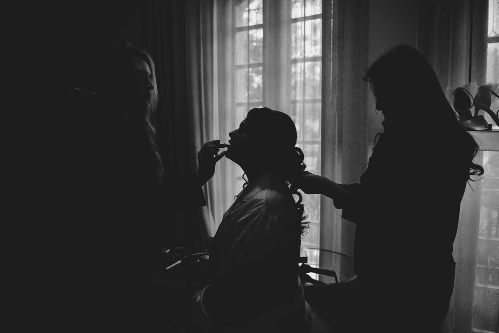 Silhouette of bride getting hair styled by two assistants in front of bright window, black and white wedding photo.