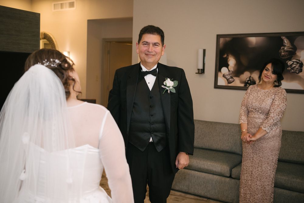 Father of bride smiling joyfully at daughter in wedding dress during first look reveal in hotel room.