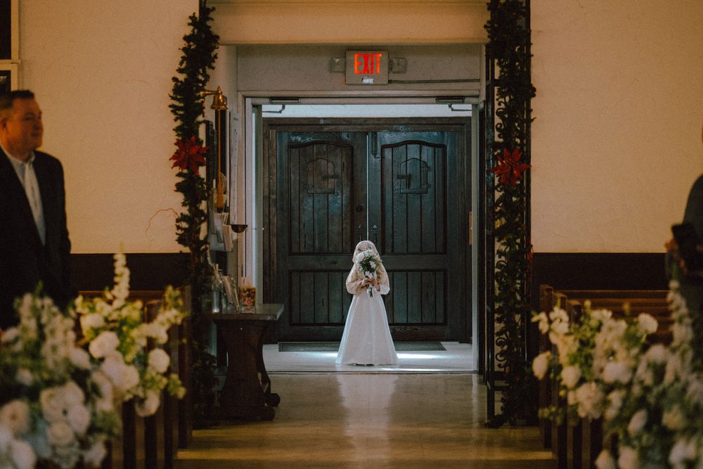 Bride in white gown and veil standing at church entrance doors lined with floral arrangements before ceremony.