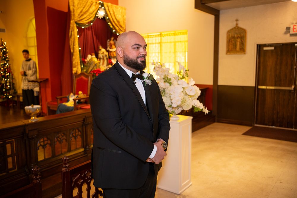 Groom in black tuxedo standing at altar with white floral arrangement in decorated church during wedding ceremony.
