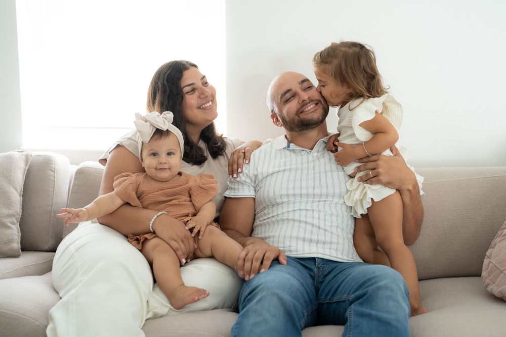 Happy family of four sitting on a cozy sofa, with parents holding two young daughters in a bright living room.