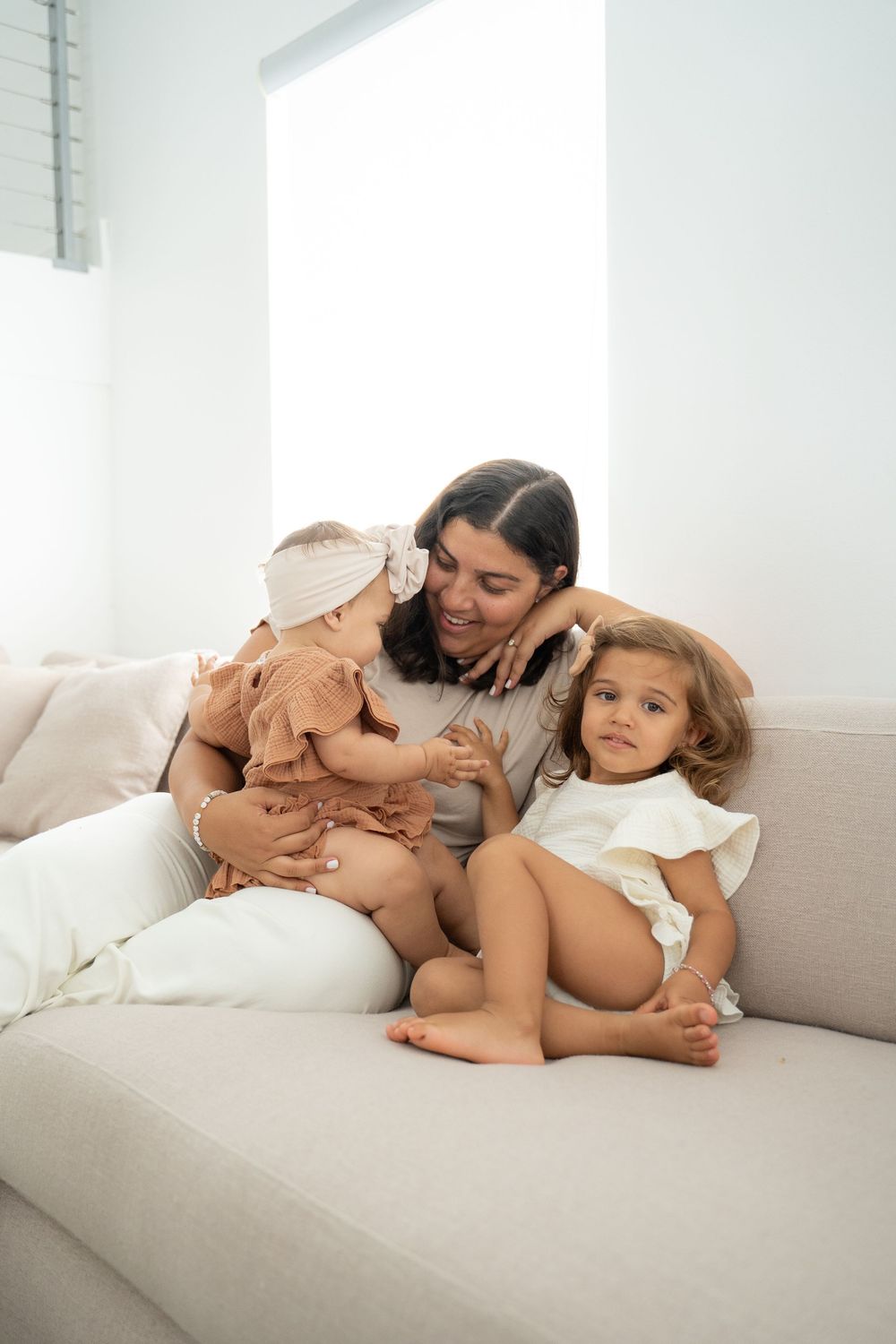 A mother sits on a beige sofa smiling at her two young daughters in a bright, airy living room.