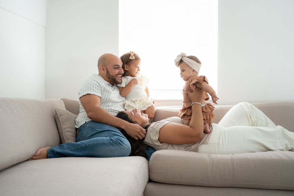 Happy family with two young daughters relaxing together on a light gray sectional sofa in a bright living room.