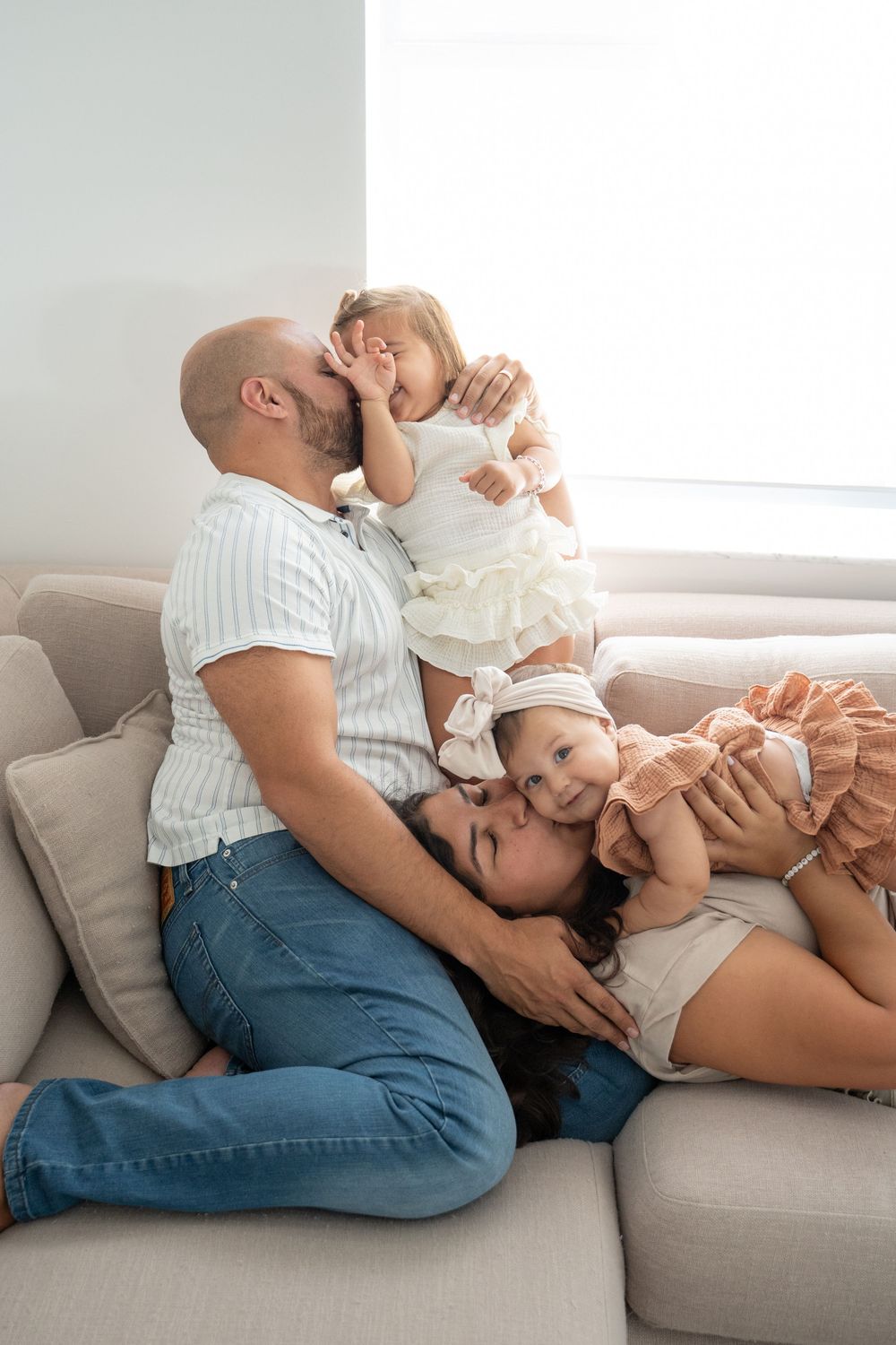 Happy family with two young daughters cuddling on a beige sofa in a bright living room.