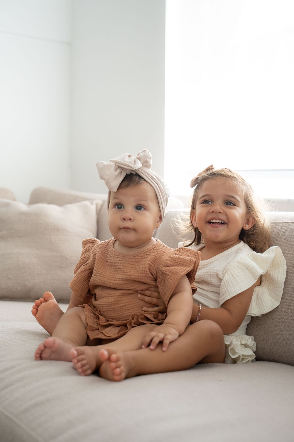 Two adorable baby girls in ruffled outfits and bow headbands sitting together on a neutral couch, smiling cutely.