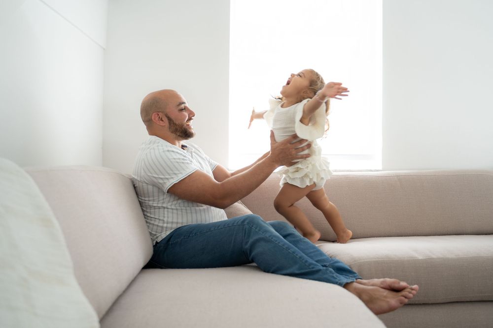 Smiling bald father lifting happy baby girl on beige sofa in bright white living room.