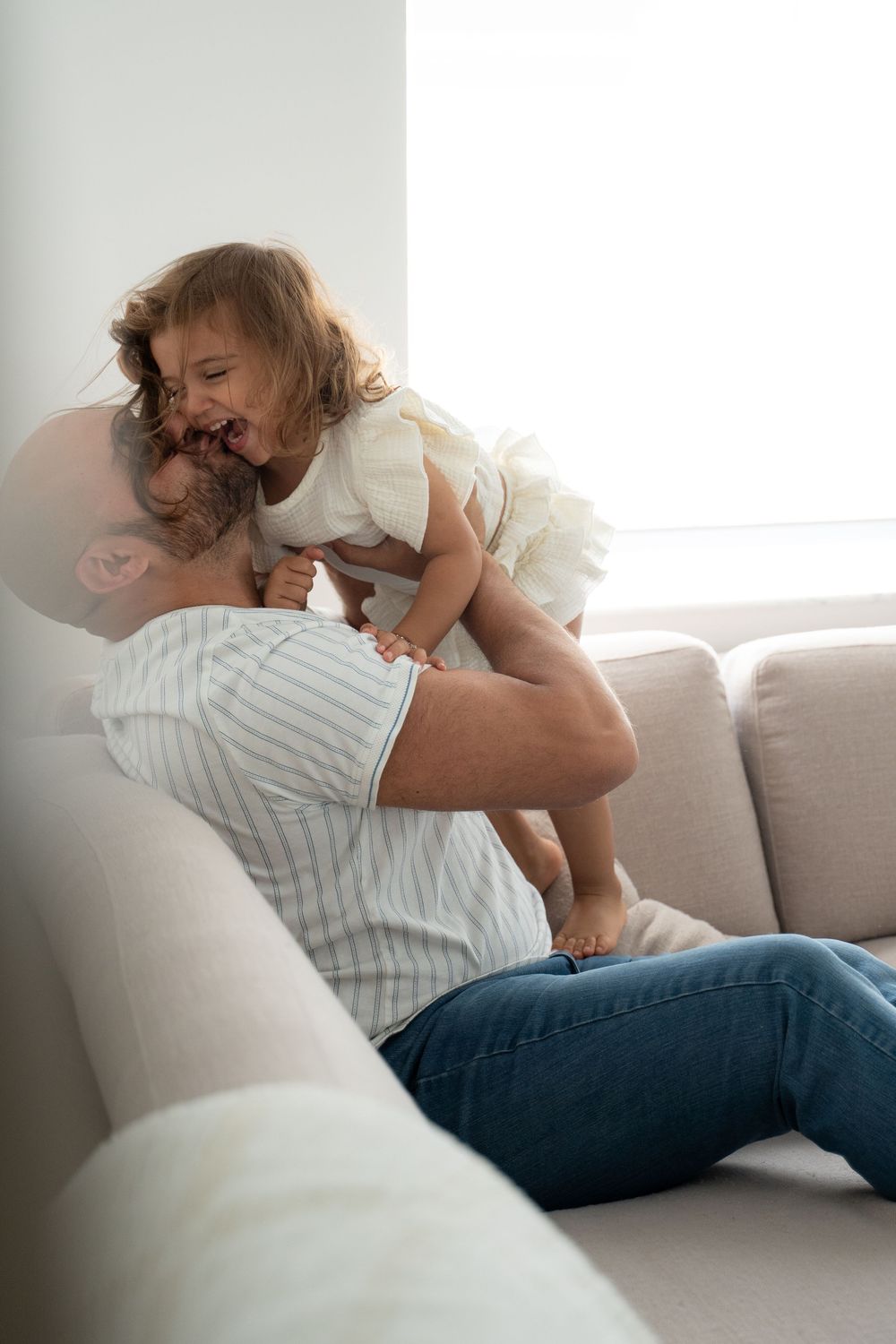 A father playfully lifts his laughing toddler daughter on a couch in a bright living room.