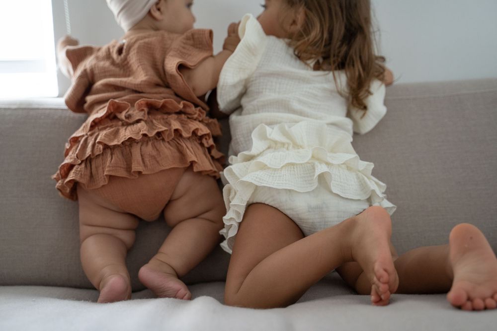Two baby girls in ruffled bloomers sitting on a grey sofa, wearing brown and cream outfits.