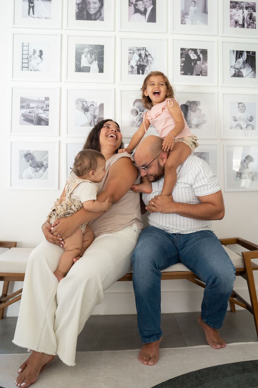 Happy family of four laughing together on a bench, with a gallery wall of black and white photos behind them.