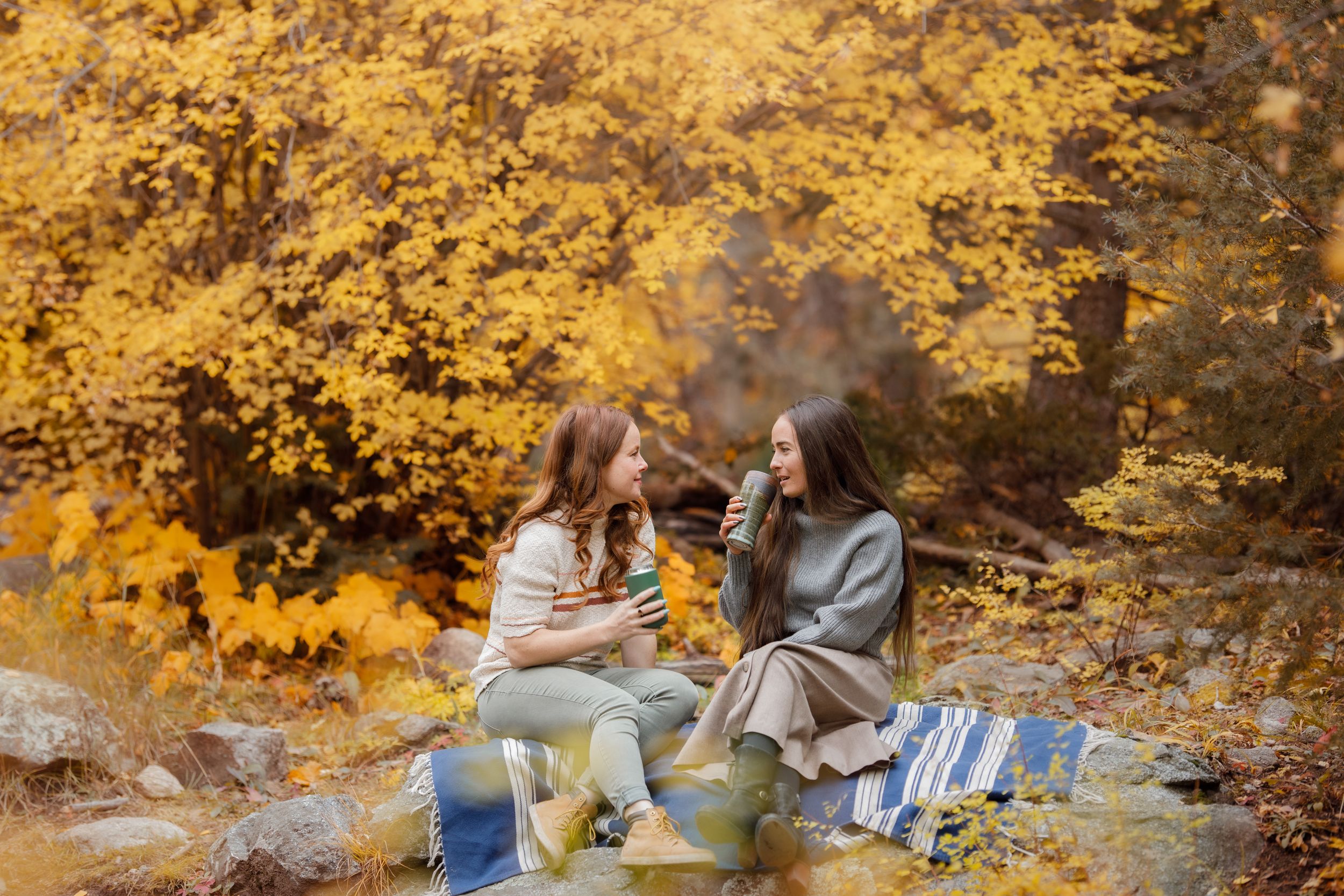 Two women enjoy hot drinks on a blanket surrounded by vibrant golden autumn foliage in a forest setting.