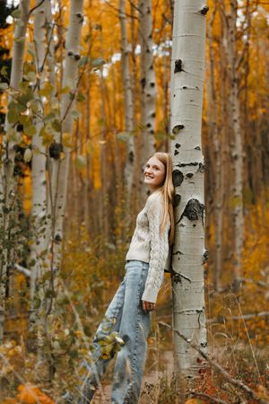 A smiling girl in a cream sweater and jeans leans against a white birch tree in a golden autumn aspen forest.