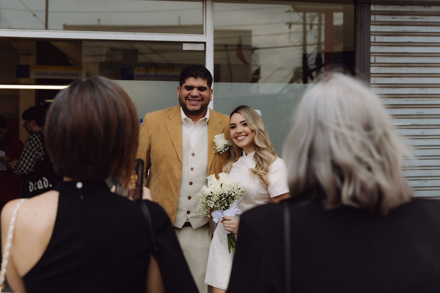 Newlywed couple smiling at guests outside a building, bride holding white bouquet and groom in mustard jacket.