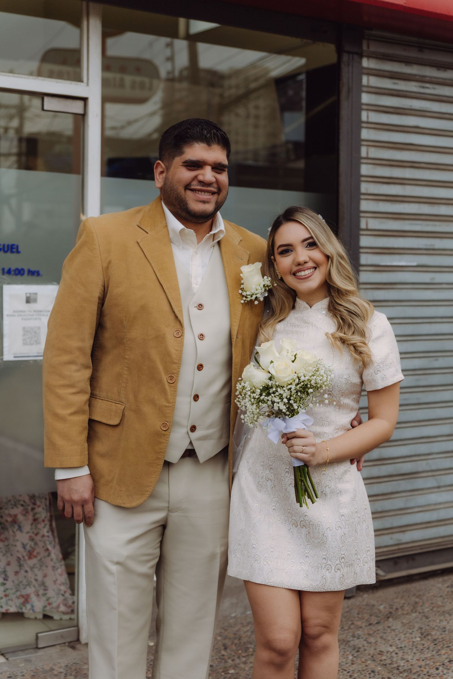 Smiling newlywed couple posing outside a building, bride holding white bouquet in short dress and groom in mustard blazer.