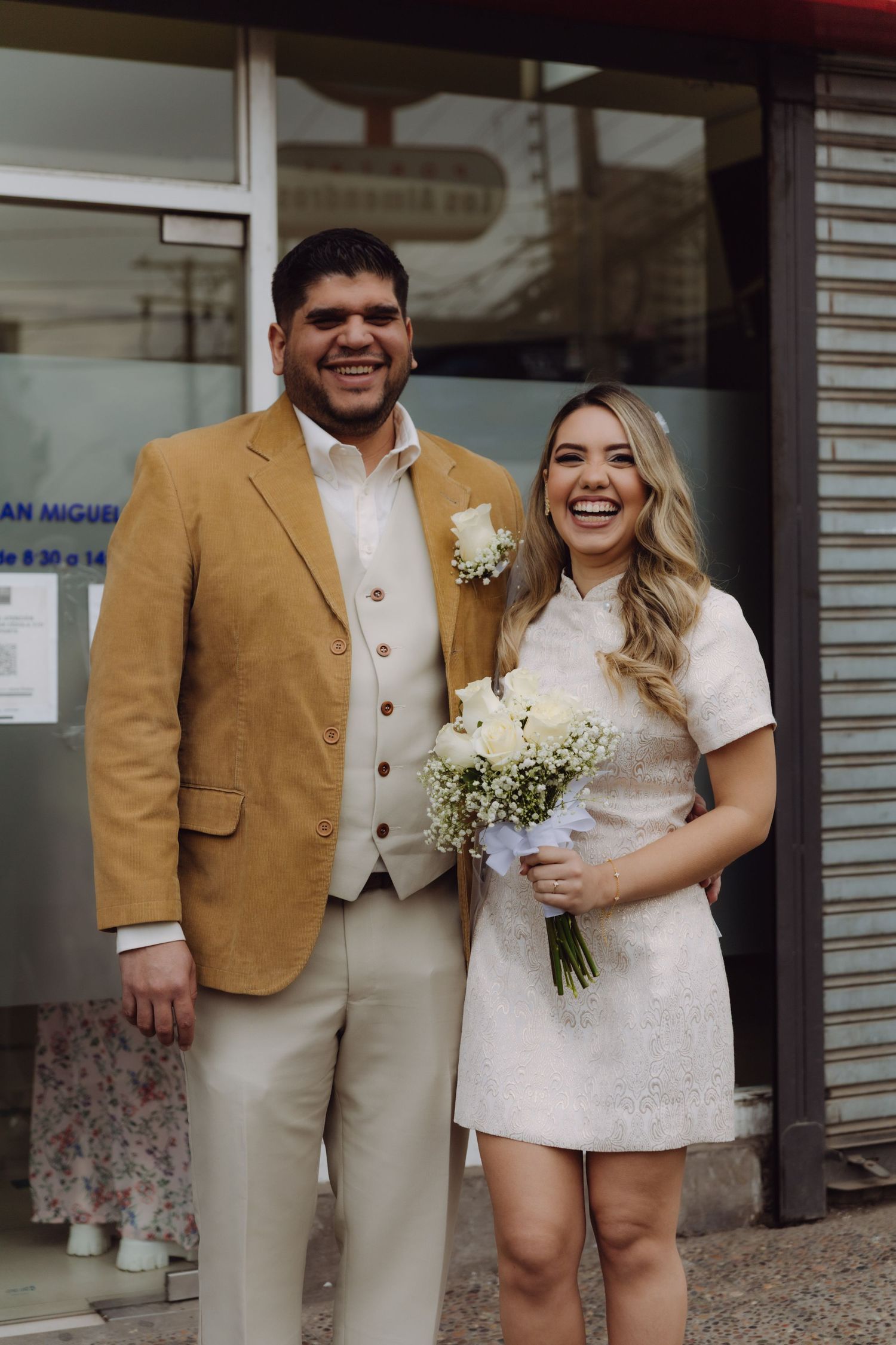 Laughing newlywed couple posing outside after their ceremony, bride with white bouquet and groom in mustard jacket.