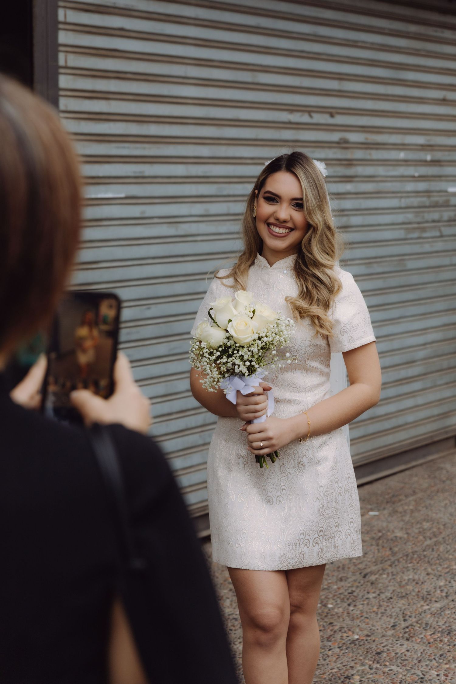 Smiling bride in white mini dress holding bouquet of white roses while friend photographs her outdoors.