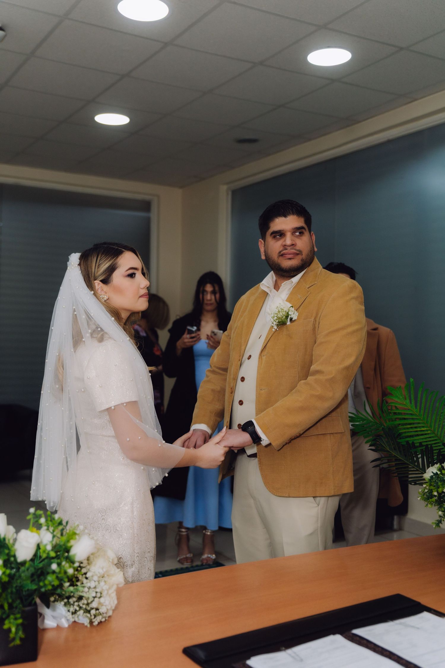 Bride in white lace dress and veil holds hands with groom in tan blazer during indoor wedding ceremony.
