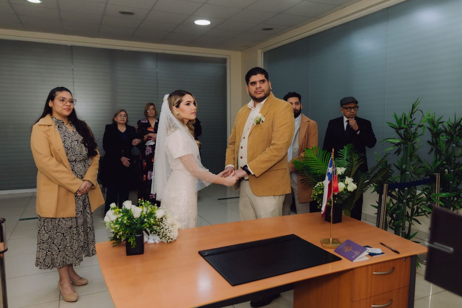 Bride and groom hold hands at wedding ceremony desk surrounded by family and friends in indoor venue.