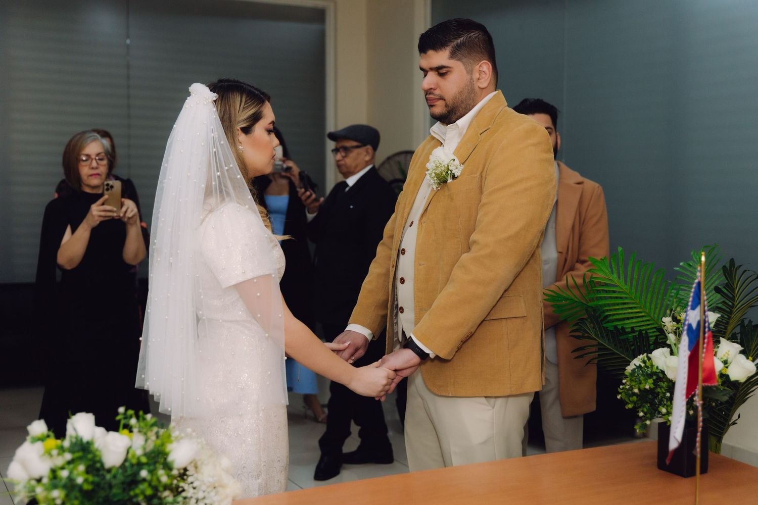 Bride in white veil and groom in mustard blazer hold hands during intimate civil wedding ceremony.