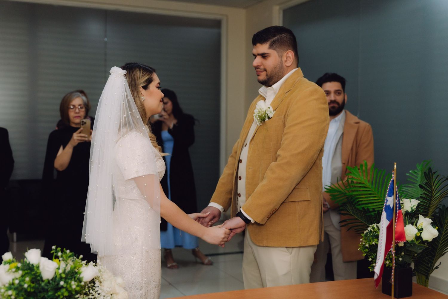 Bride and groom face each other holding hands during wedding ceremony with guests watching in background.