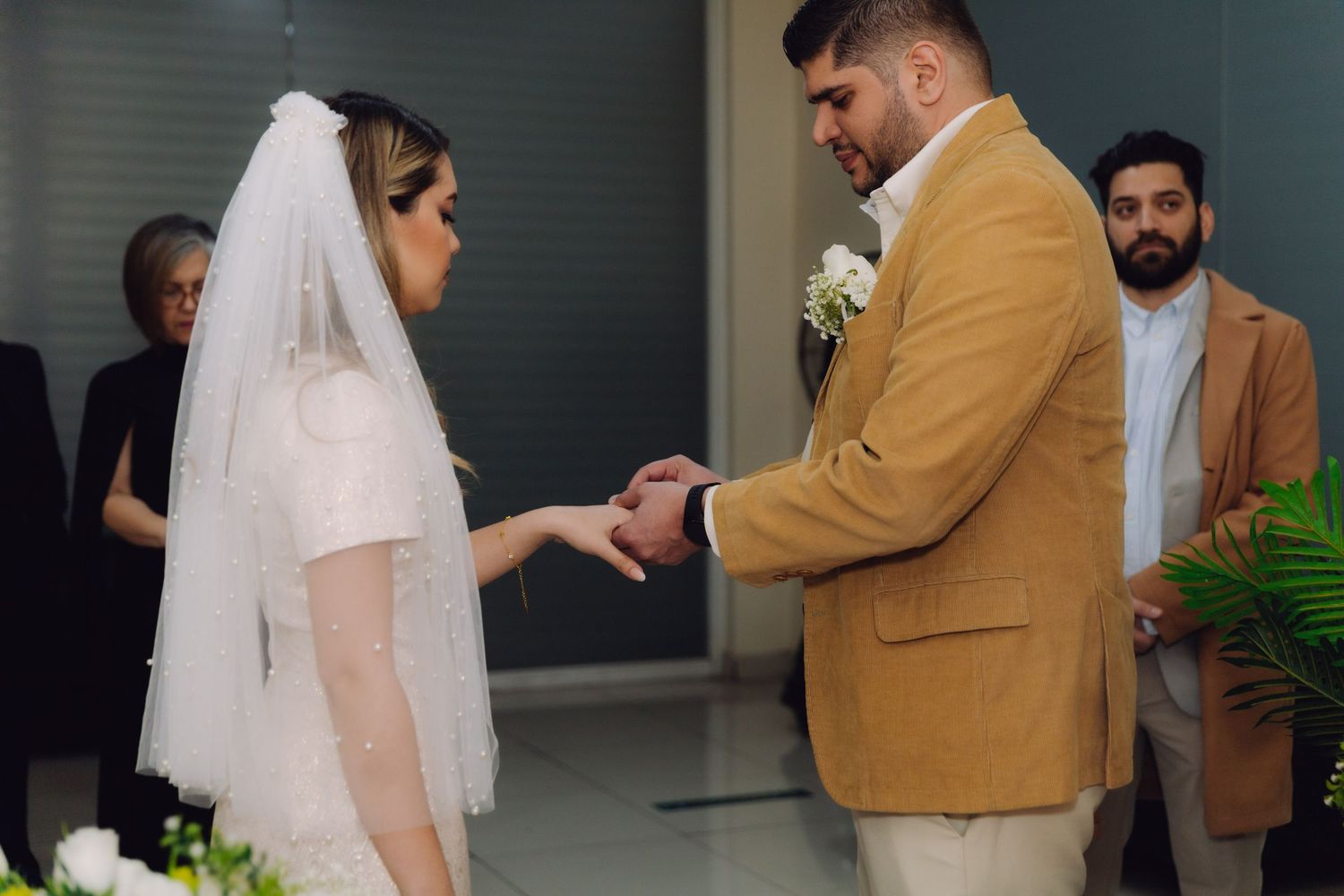 Groom places wedding ring on brides finger during civil ceremony with white floral arrangements nearby.