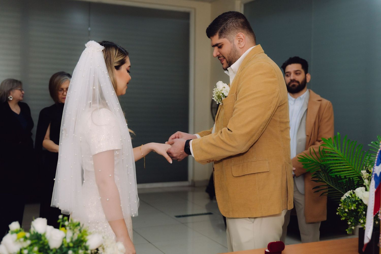Groom slides wedding ring onto brides hand during indoor civil ceremony with guests in background.