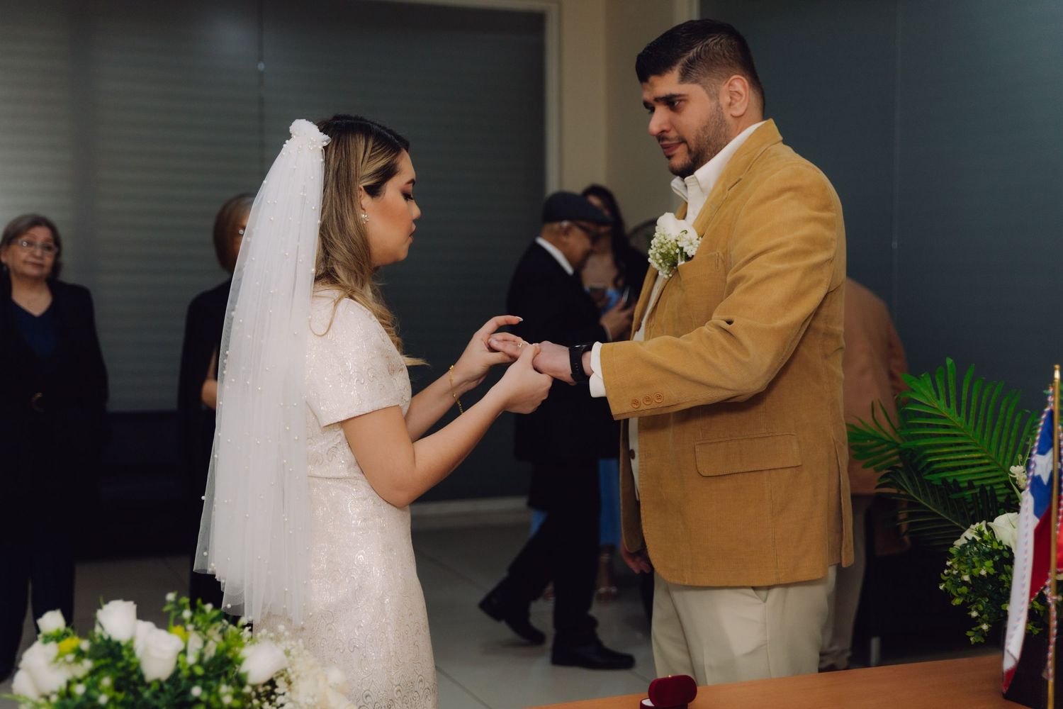 Bride places wedding ring on grooms finger during civil ceremony with white flowers and flag nearby.
