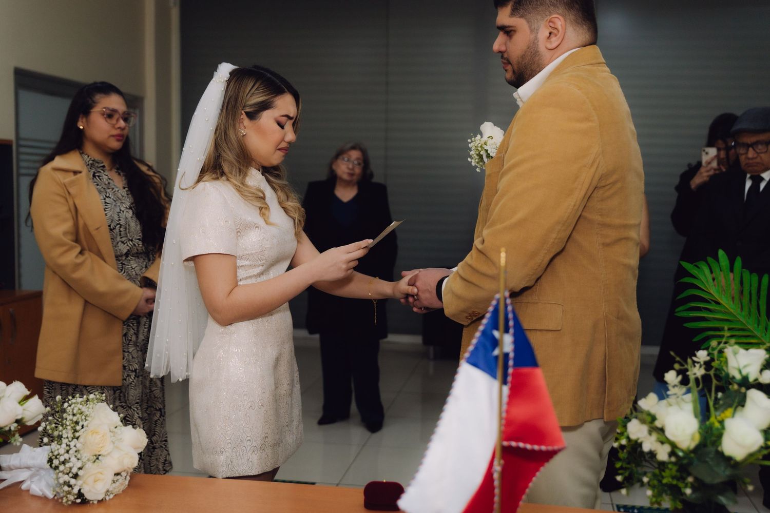 Bride reads vows to groom with Chilean flag visible during civil wedding ceremony.