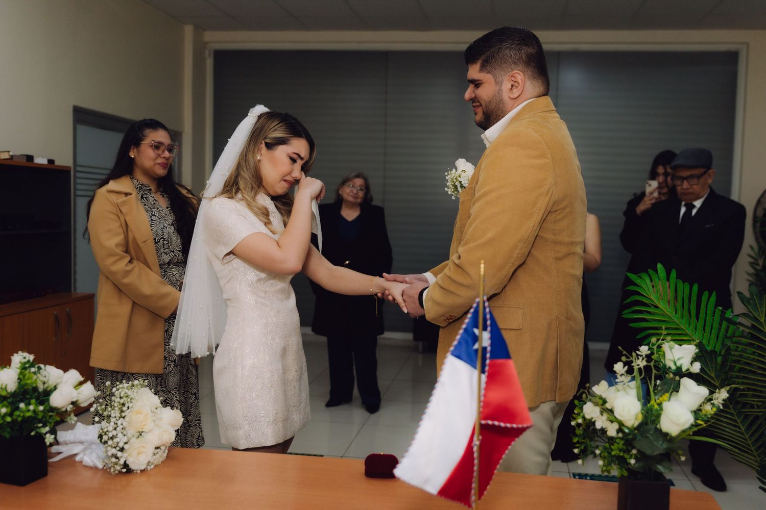 Emotional bride wipes tear while holding groom's hands at civil wedding ceremony.
