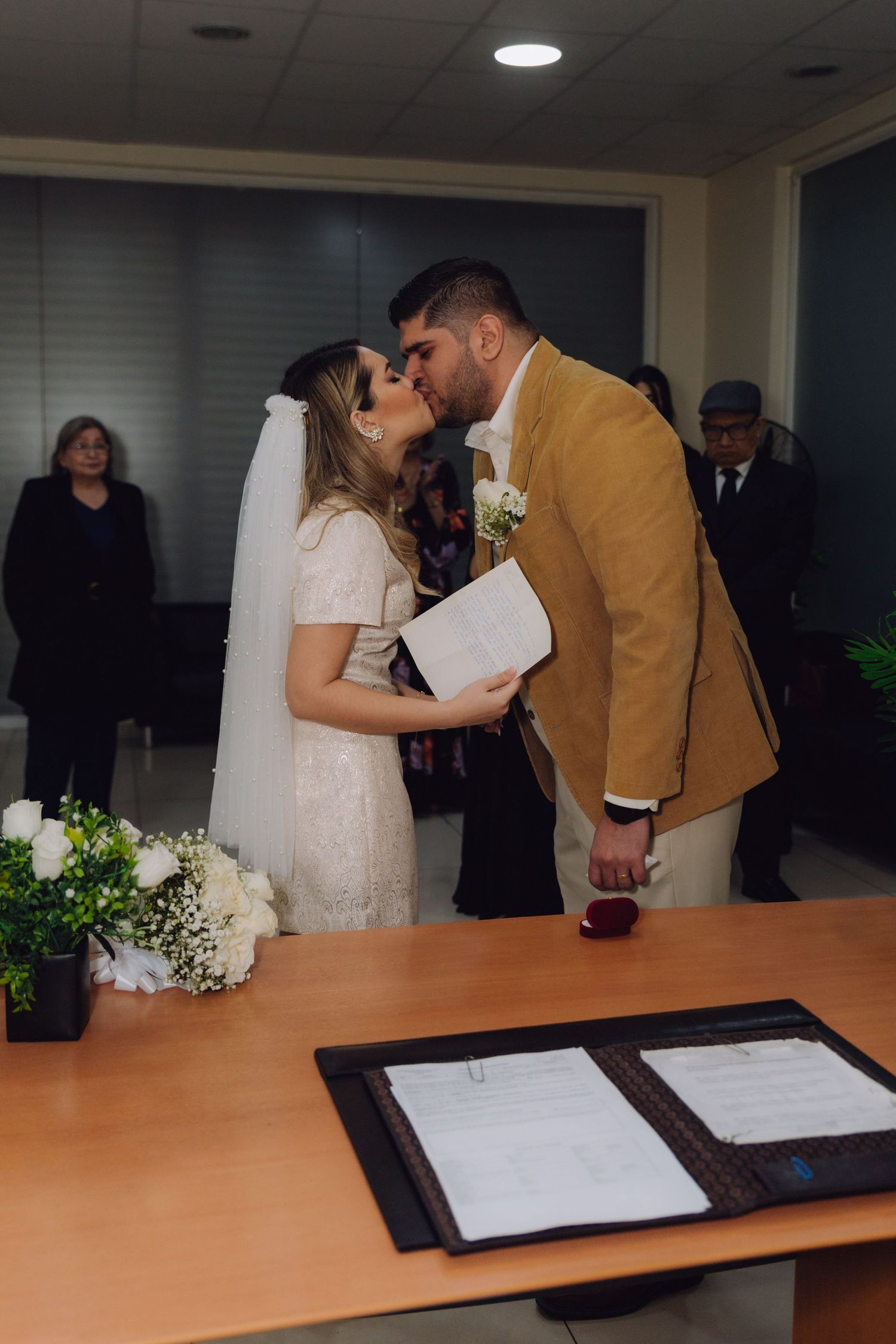 Newlyweds share first kiss at civil wedding ceremony with guests watching in background.