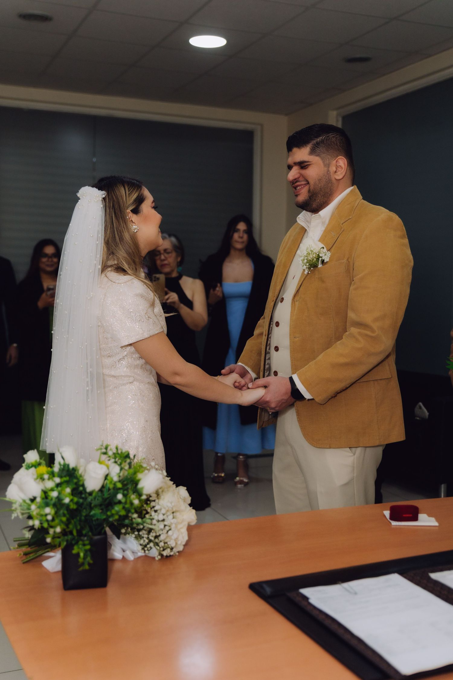Smiling groom holds bride's hands during joyful civil wedding ceremony with guests behind them.
