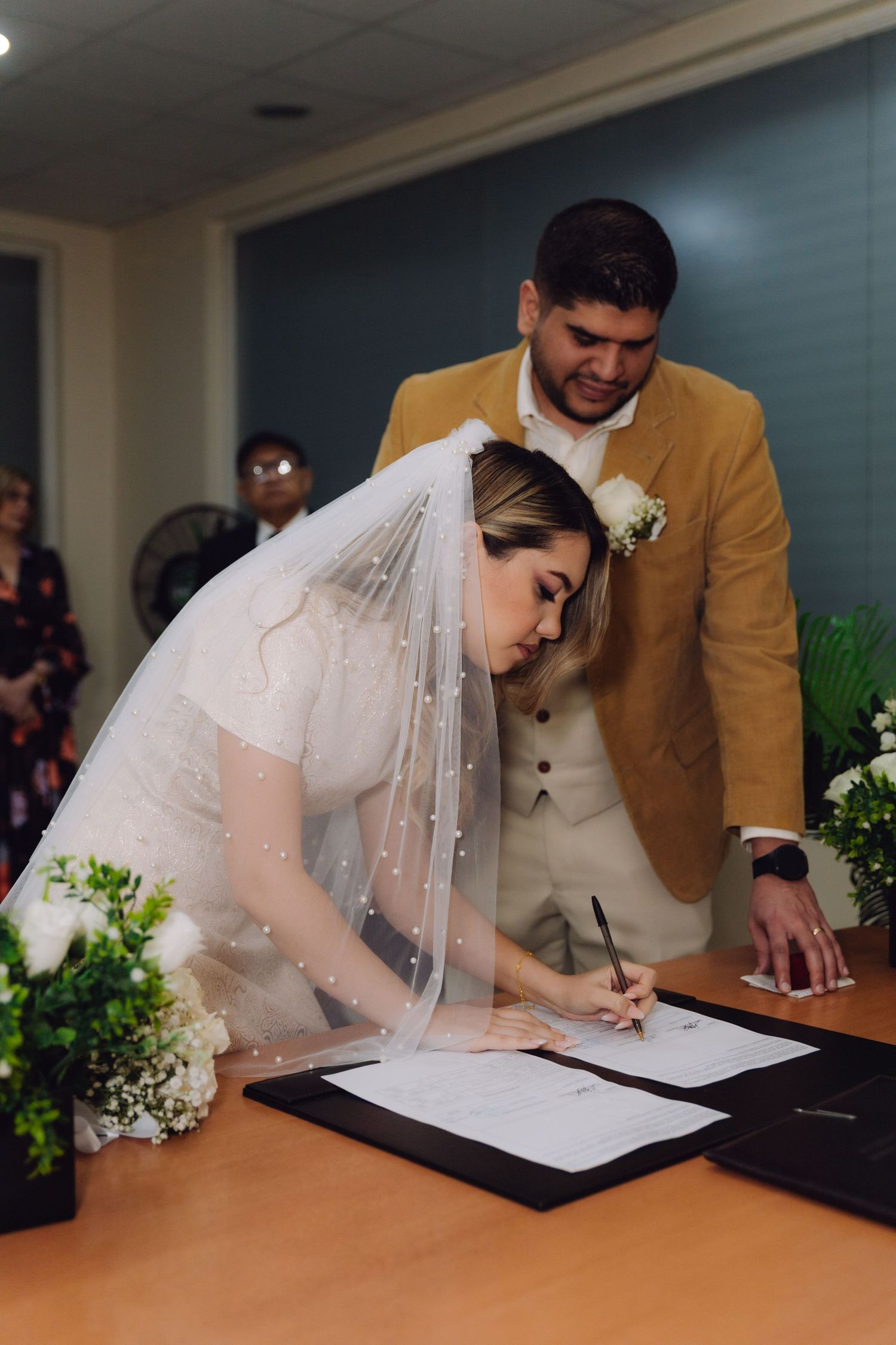 Bride signing wedding documents while groom in mustard suit stands beside her.