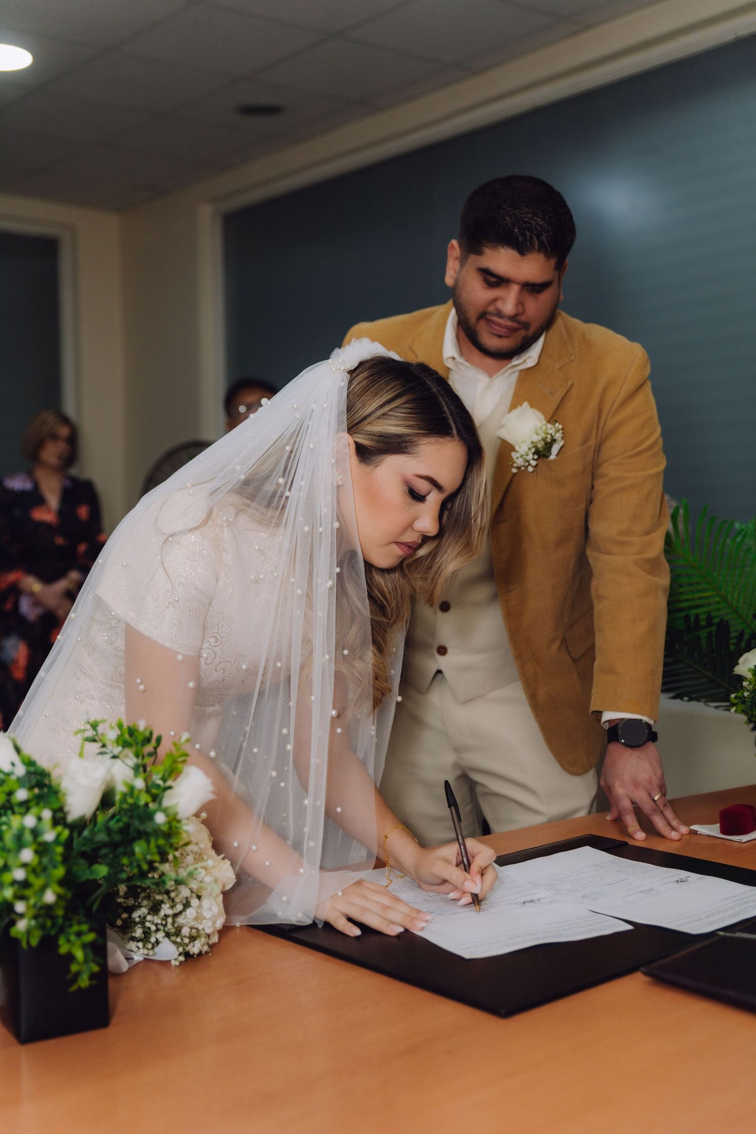 Bride in white lace dress and veil signing marriage certificate as groom watches.