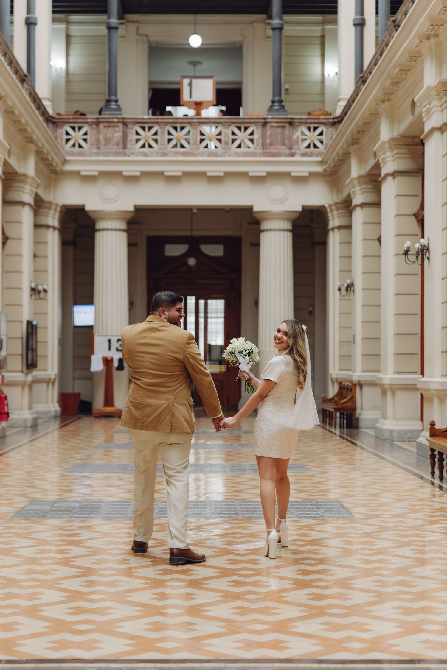 Bride and groom holding hands in an elegant historic building with ornate columns and decorative tiled floors.