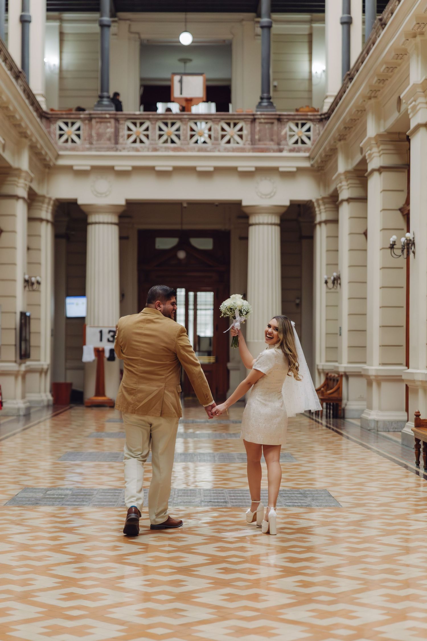 Wedding couple walking hand in hand through a grand colonial-style hall, bride holding white bouquet and veil.