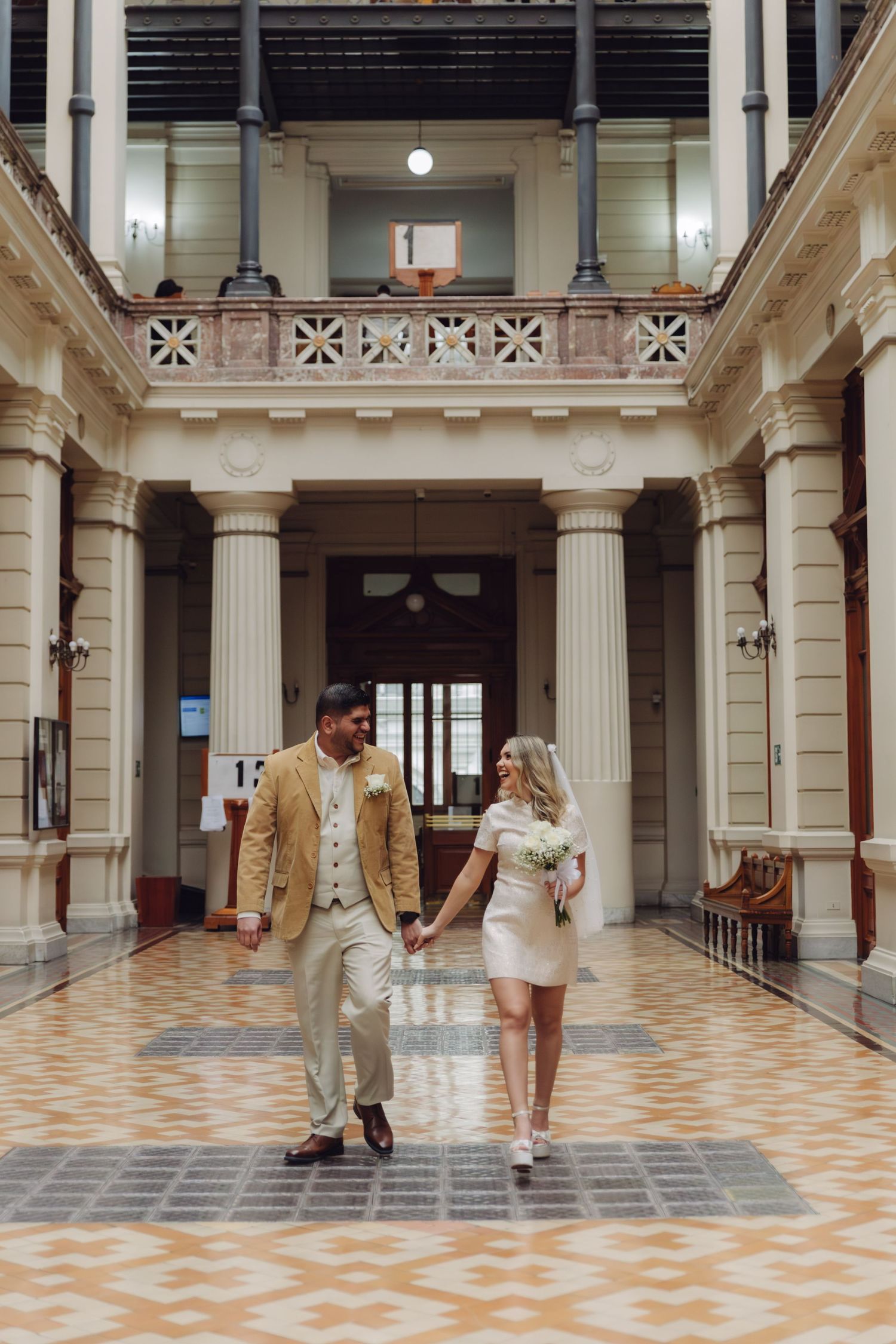 Newlyweds strolling together through a stunning architectural corridor with tall white columns and patterned floors.